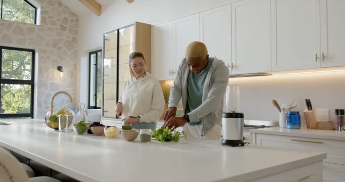 Seeing ingredients, Diverse couple at kitchen island, chopping herbs and slicing cucumber for salad