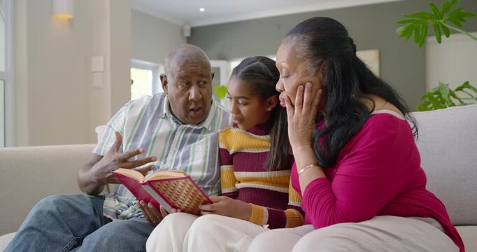 African American grandparents with youth girl sitting on sofa reading book gran pointing and miming