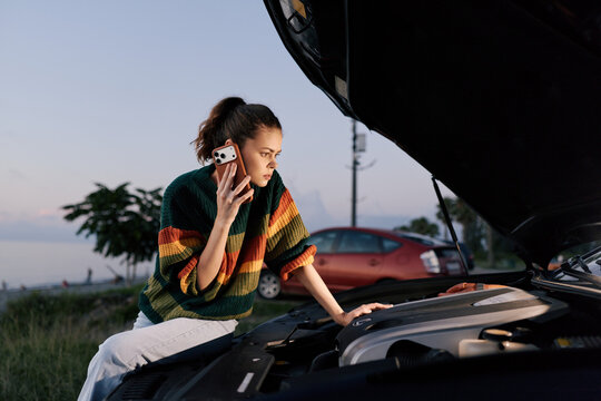 Young woman calls for roadside assistance while looking at broken car engine with open hood outdoors. Stressful situation during travel emergency help.