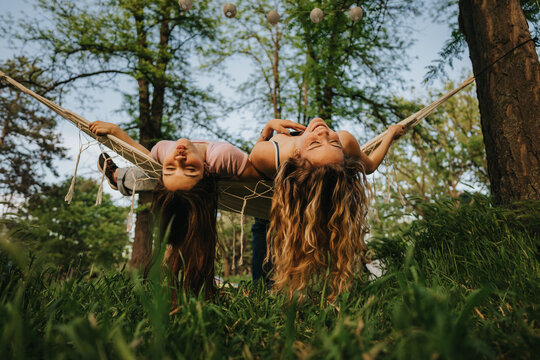 Two teenage friends lie upside down on a hammock in a leafy park, smiling and laughing. They relax together outdoors, enjoying a carefree summer moment amid trees and grass.