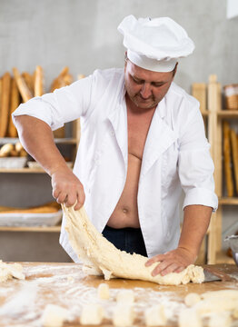 Male baker in unbuttoned uniform prepares dough, kneads it with his hands