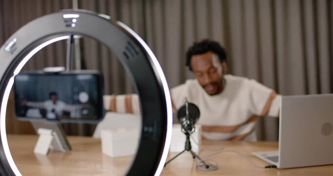 African American man hosting stream at desk under ring light, phone LED signaling gesturing to mic