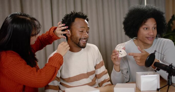 African American co-hosts inserting earbud into center man, showing case while testing fit