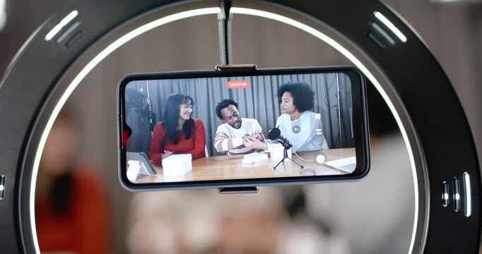Diverse co-hosts recording in studio red REC flashing on phone in ring light, reviewing smartwatch