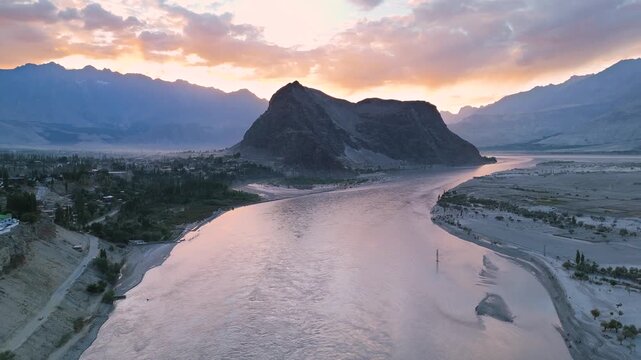 4K drone orbit shot of the Indus River flowing past a majestic mountain in Skardu during a vibrant sunset, panoramic view of a rocky peak and wide glacial river bathed in pastel evening light