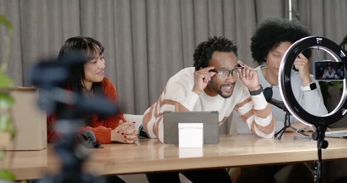 Three hosts in studio ring light, right tapping phone, center testing eyeglasses demoing features