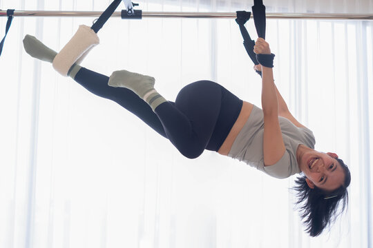 Woman smiling while performing aerial Pilates with suspension straps in a bright fitness studio, demonstrating strength, flexibility, balance, core control, mobility, and active wellness.