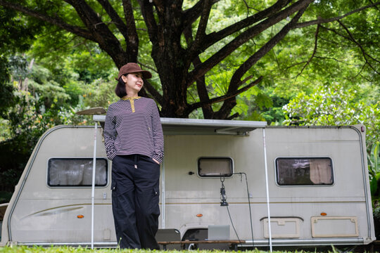 Woman relaxing beside a camper trailer in a lush forest, enjoying outdoor travel, camping, nature, freedom, adventure, peaceful recreation, and a flexible lifestyle surrounded by greenery.