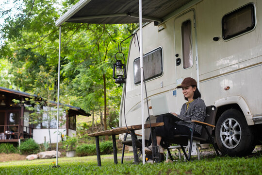 Woman working on a laptop beside a camper trailer at a forest campground, combining remote work, camping, outdoor travel, digital nomad lifestyle, relaxation, nature, and work-life balance.