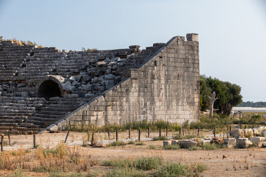 A historic stone amphitheater ruins with terraced seating, an arched entrance, and weathered walls sits in a sunlit landscape. Roman Theatre of Patara ancient city, Turkey
