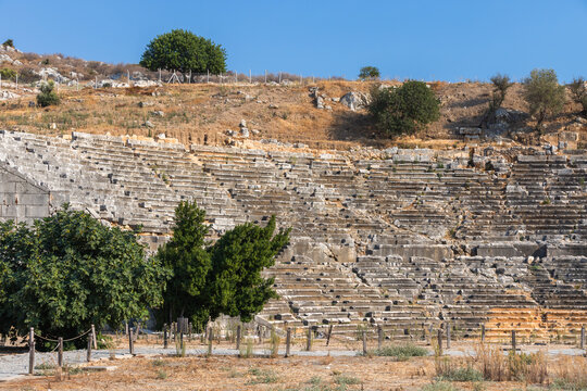 A historic stone amphitheater ruins with terraced seating, an arched entrance, and weathered walls sits in a sunlit landscape. Ruined Roman Theatre of Patara, Turkey