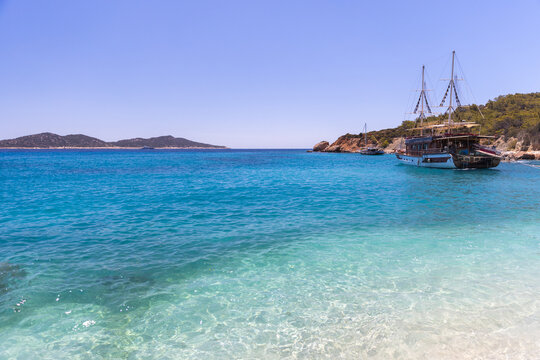 Blue sea laps a sandy shore beside rocky cliffs where are anchored sailings. Bright blue sky and clear water evoke summer travel, relaxation, coastal adventure and seascape beauty. Turkey