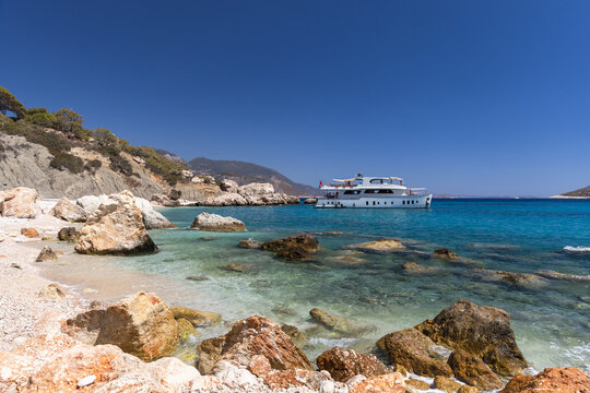 Tourist boat is near a rocky Turkish beach with crystal turquoise water, pebble shoreline and distant hills under a clear blue sky, evoking summer travel, relaxation and coastal adventure