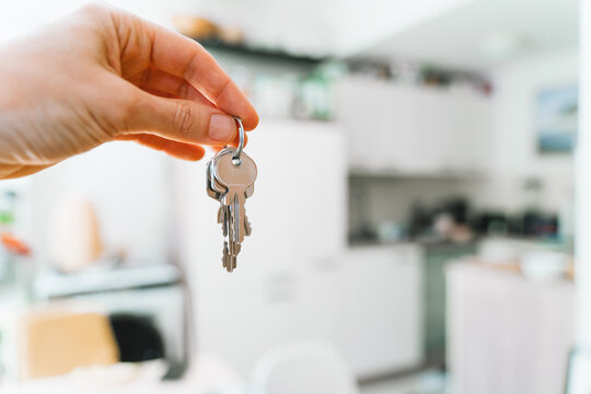 A hand holds new house keys against the blurred background of a bright modern apartment kitchen. Successful residential property purchase and exciting relocation commercial lifestyle concept.
