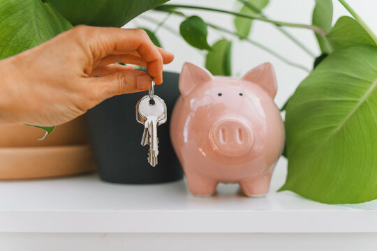 A female hand dangles a house key next to a pink piggy bank and a green potted plant on a shelf. Financial savings for mortgage investment and property ownership lifestyle concept.