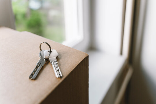 A shiny set of house keys lies abandoned on a cardboard moving box right next to a bright window. Authentic property ownership and exciting relocation transition lifestyle concept.