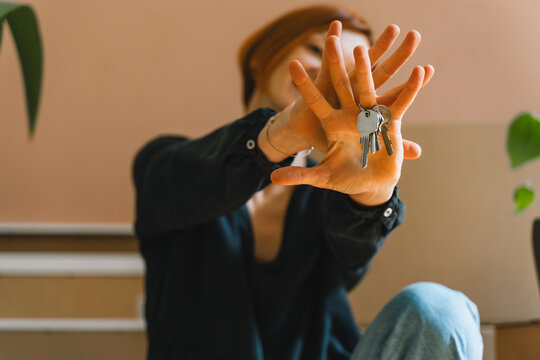 A happy Caucasian woman sits among cardboard boxes and playfully shows her new house keys to the camera. Authentic real estate ownership and successful relocation lifestyle concept.