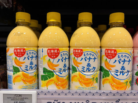 Four bottles of yellow banana milk are displayed on a store shelf. The labels feature vibrant illustrations of bananas and Japanese text. The scene showcases dairy beverages in a retail setting.