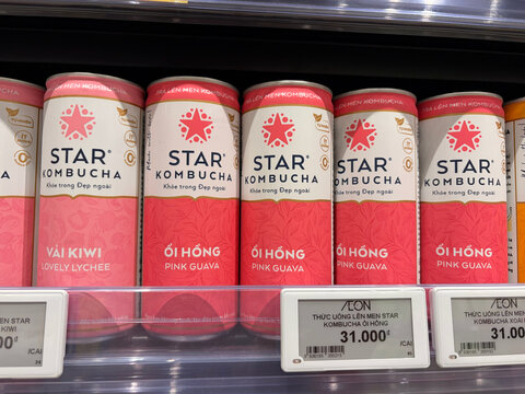 A row of cans featuring STAR Kombucha displayed on a supermarket shelf. The cans are vibrant pink and white, labeled with flavors. Price tags are visible beneath each can.