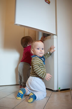 Curious twins babies exploring the white kitchen refrigerator during a quiet afternoon at home while searching for snacks in their gentle household