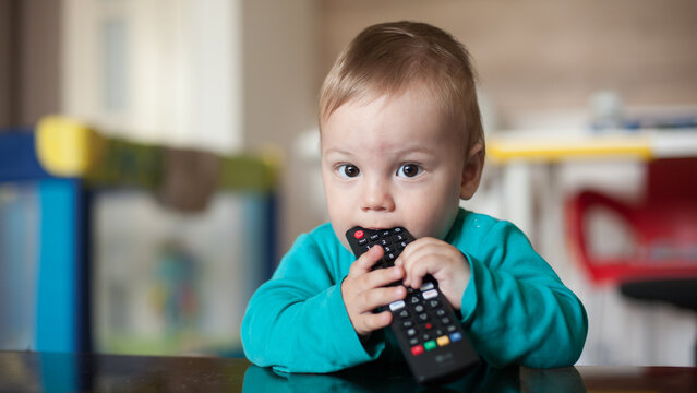Small baby boy sits at a table and curiously bites down on a black television remote control while holding the device with both tiny hands indoors