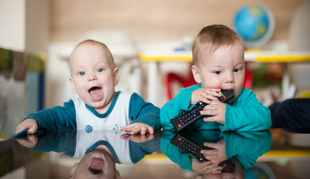 Two toddler twins baby brothers share a joyful moment at home as one chews a remote and the other laughs with excitement
