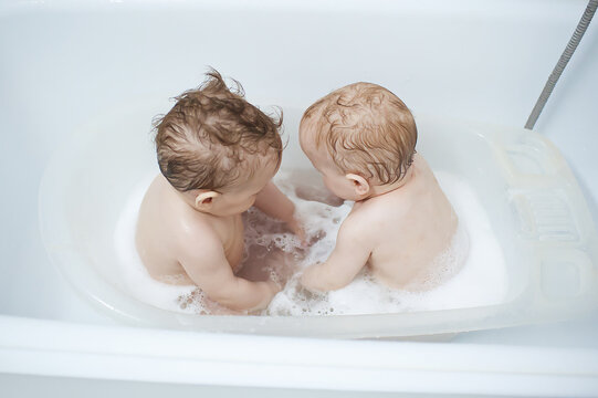 Twins babies splashing in a small white bathtub filled with bubbly water and soap during a gentle evening hygiene ritual in the bathroom