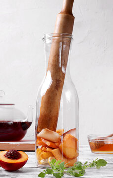 Nectarine and lemon pieces and a wooden muddler in a glass bottle on a white table. Arranged around it are black tea in a glass teapot, honey and mint sprigs. Making iced fruit tea. Step-by-step
