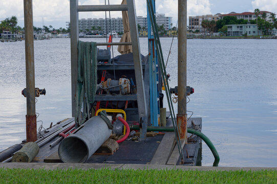 Cluttered marine work dock with hoses, pipes, ladder and winch on a small pier beside calm harbor water and distant waterfront buildings, suggesting industrial maintenance and coastal utility operatio