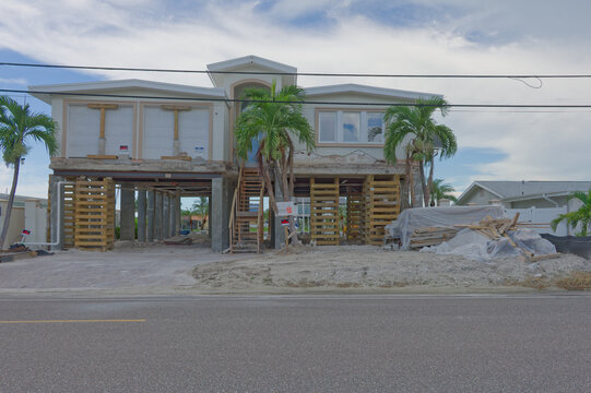 A coastal two-story house raised on concrete pilings and temporary wooden supports during renovation after storm damage, with palm trees, construction materials and an access stairway visible from the