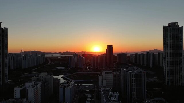 4K aerial drone footage flying past tall residential towers as the sun sets behind the Incheon skyline in South Korea. Foreground skyscrapers move against the distant city, and the backlit buildings b
