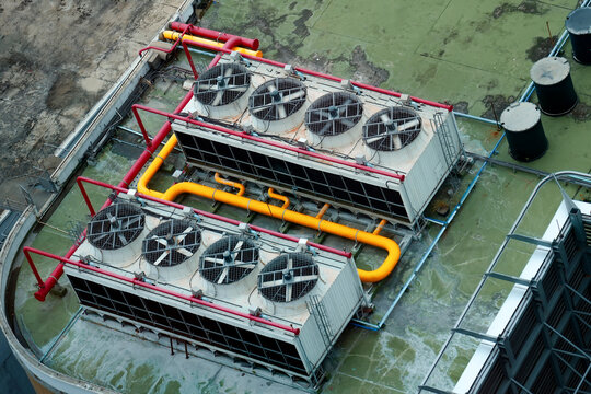 Massive industrial cooling towers stand on a rooftop with bright yellow and red pipes connecting the metal machinery under the clear daylight sun