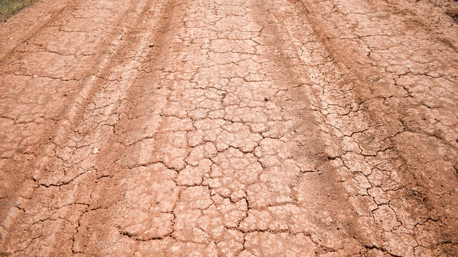 Detailed close-up shot showing the texture of parched, dry red earth on a rural track. Deep tire tread marks are clearly visible on the cracked surface under bright natural daylight