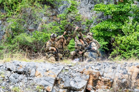 Group of soldiers in tactical gear training in a rocky area surrounded by green foliage during the day