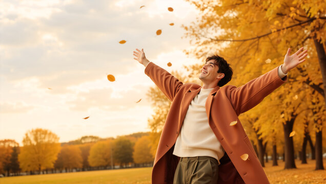 Smiling young man standing in an autumn park with arms outstretched. Happy male enjoying falling yellow leaves in nature. Freedom and vitality lifestyle concept