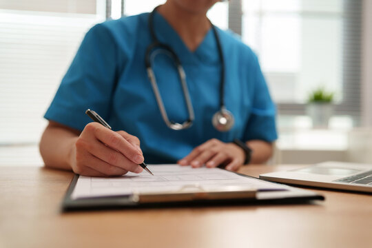 Doctor in blue scrub uniform writing and reviewing medical documents at a clinic with laptop, representing healthcare records, patient examination, diagnosis and professional medical care.