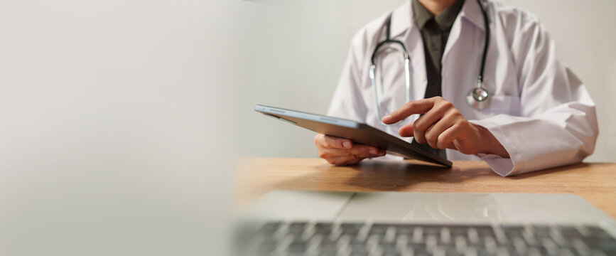 Doctor using digital tablet and laptop at a medical desk, representing healthcare technology, patient diagnosis, clinical review, digital records and modern professional medical care.