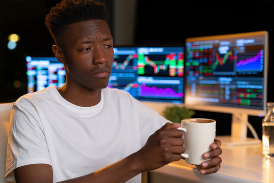 Young African American man in white t-shirt holding coffee mug while sitting at desk with multiple computer screens displaying financial charts and data at night