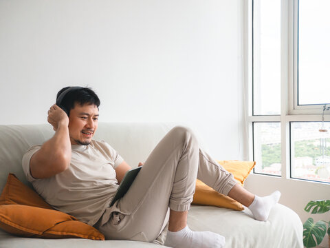 Happy Asian man enjoying leisure time, watching content on tablet with headphones at home.