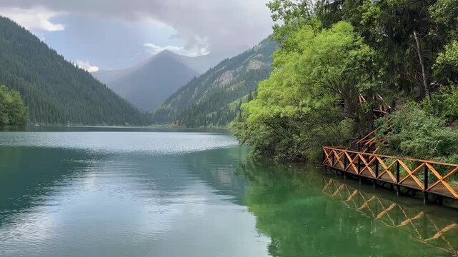 Scenic Mountain Lake in Kazakhstan Surrounded by Dense Forest and Tien Shan Mountains with Wooden Lakeside Walkway on an Overcast Day
