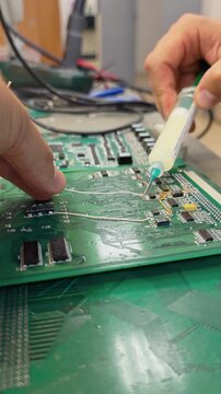 Hand holding a flux syringe over a green circuit board during component replacement, close-up electronic repair, precise soldering work.