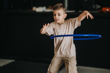 A young boy plays with a blue hula hoop indoors, concentrating on keeping it spinning. Focused child practices balance and movement while enjoying a simple active toy. © qunica.com