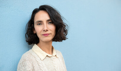 Woman with short wavy bob stands against a blue wall while looking at the camera in a casual setting © nikopacheco