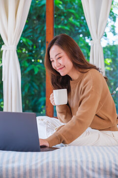Vertical portrait of a woman using and working on laptop computer while sitting and drinking coffee on the bed