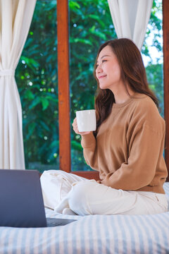 Vertical portrait of a woman using and working on laptop computer while sitting and drinking coffee on the bed