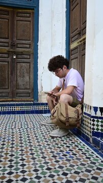 Marrakech, morocco. August 10, 2026. Young man with curly hair and glasses sitting on traditional moroccan tiles in a courtyard, focused on his mobile phone in marrakech