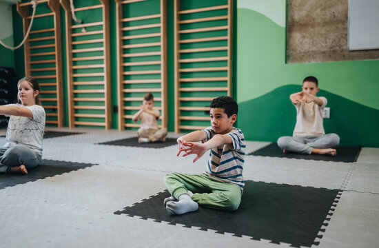 A group of children sit on mats stretching their arms during a supervised gym session. Young students practice seated exercises and calm breathing in a school fitness class.