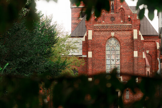 Gothic red brick cathedral facade framed by lush green foliage under soft natural light glare
