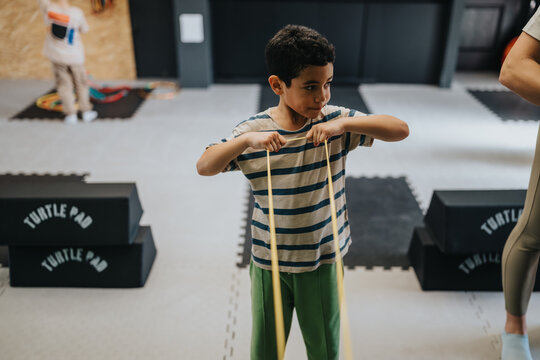 A young boy stretches a yellow resistance band while standing in an indoor kids gym. The child practices coordination and balance among foam mats and exercise equipment.