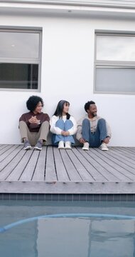 Vertical video: Diverse friends sitting on deck above pool center glancing causing man laughing